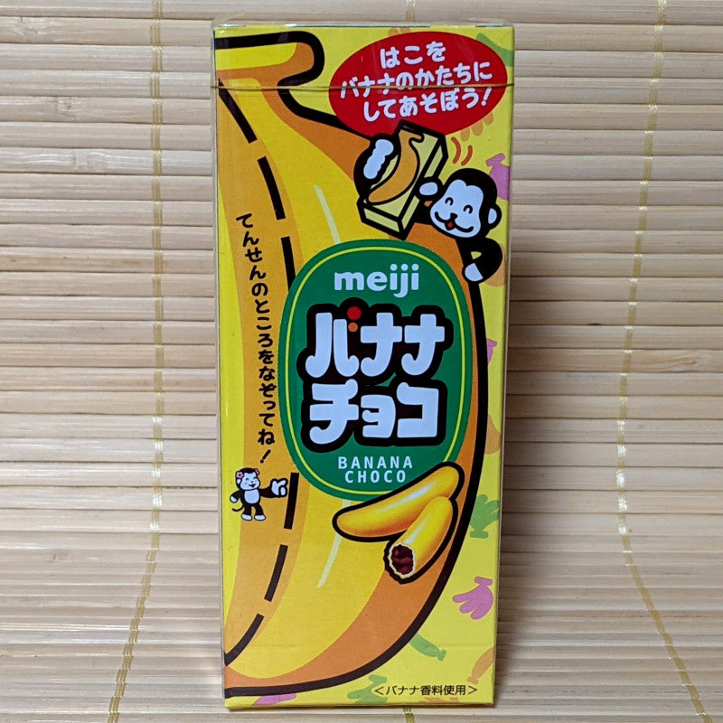 A package of Meiji Banana Choco, which are banana-shaped chocolates with crispy yellow candy coating. The packaging is predominantly yellow with illustrations of bananas and the Meiji logo.