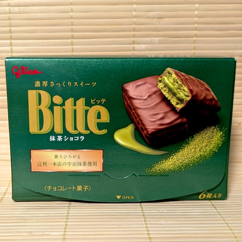 A deep green box of Glico Bitte Matcha Chocolat showing chocolate-covered biscuits with bright green matcha cream centers on a bamboo mat background.