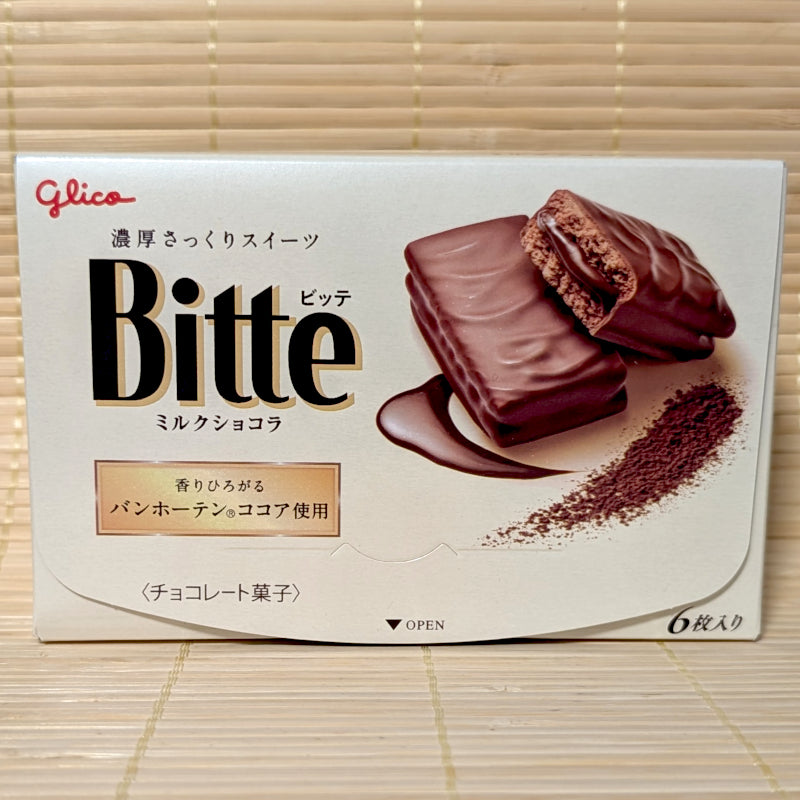 An ivory box of Glico Bitte Milk Chocolat showing chocolate-covered biscuits with creamy chocolate centers and cocoa powder on a bamboo mat background.