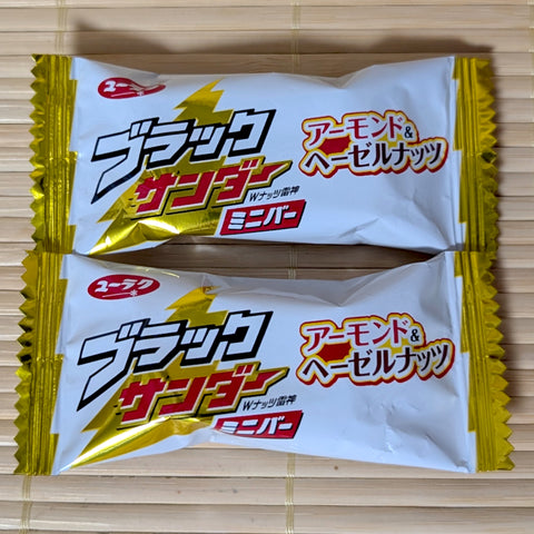 Two white and gold wrapped mini bars of Yuraku Black Thunder Almond and Hazelnut chocolate on a tatami background.