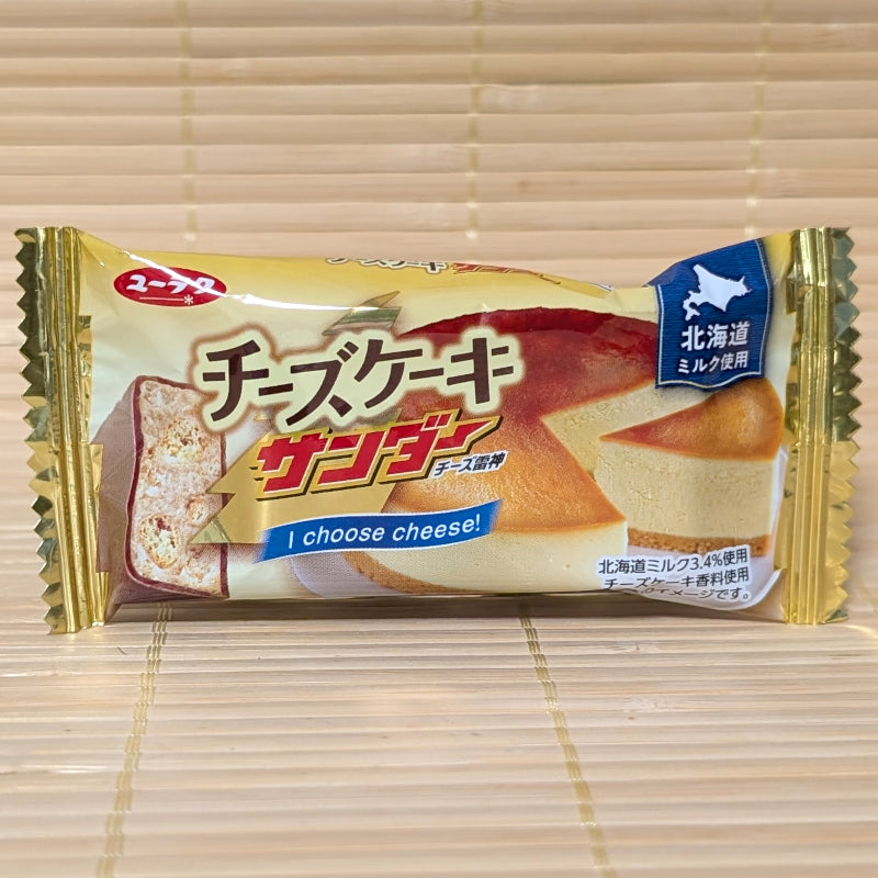 A gold and cream wrapper of Black Thunder Cheesecake edition showing a slice of baked cheesecake and a cross-section of the bar on a tatami background.