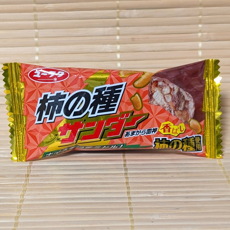 A red, gold, and yellow wrapper of Black Thunder Mini Kaki no Tane Chocolate Bar showing rice cracker graphics on a bamboo background.   