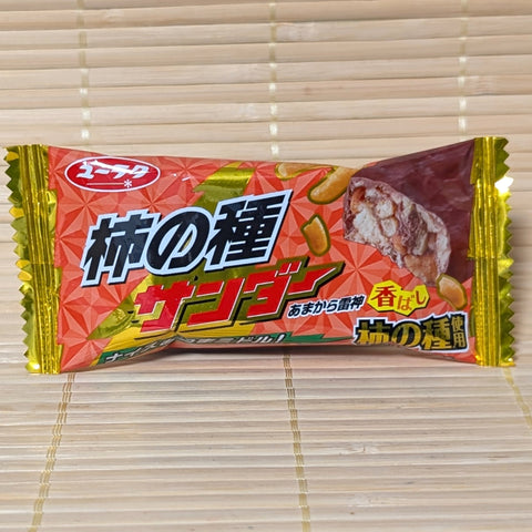 A red, gold, and yellow wrapper of Black Thunder Mini Kaki no Tane Chocolate Bar showing rice cracker graphics on a bamboo background.   