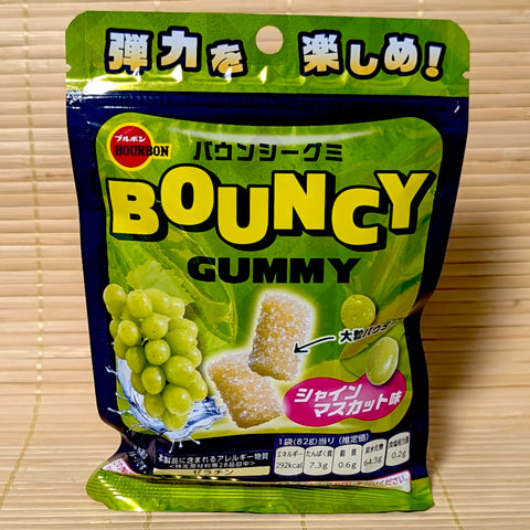 A bright green and blue pouch of Bourbon Bouncy Gummy Shine Muscat surrounded by green Shine Muscat grapes on a bamboo background.