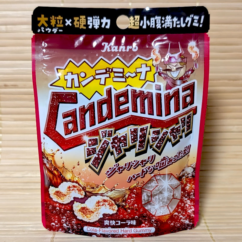A metallic red and gold 36g pouch of Kanro Candemina Jyari Shari Cola hard gummies showing a cartoon character, splashing soda, and crunchy wavy candies on a bamboo mat background.