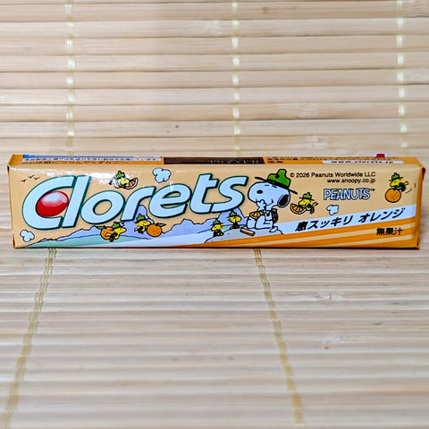 A stick pack of Clorets Fresh Breath Orange Mint Gum featuring cute Snoopy and Woodstock illustrations on a bamboo mat background.