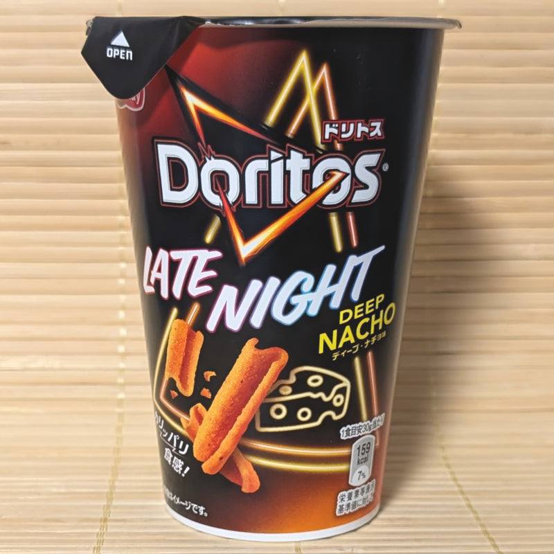A black and orange 60g snack cup of Doritos Late Night Deep Nacho showing neon lights, cheese graphics, and orange rolled tortilla sticks on a bamboo background.