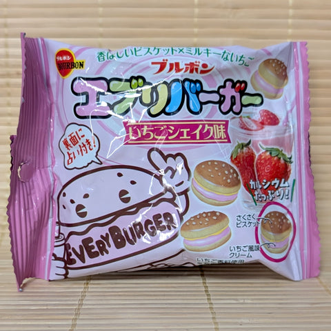 A pink 42g foil packet of Bourbon Every Burger Strawberry Shake cookies showing a cartoon burger mascot, fresh strawberries, a strawberry shake glass, and mini hamburger cookies on a bamboo mat background.