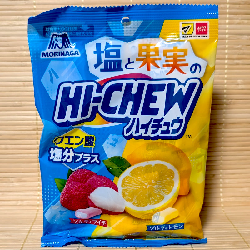 A blue and yellow split bag of Morinaga Hi-Chew Salty Lychee and Salty Lemon showing fresh sliced fruits and drugstore logos on a tatami background.