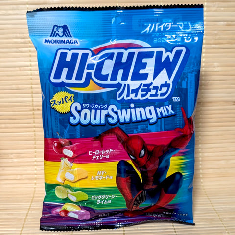 A bright blue and multi-colored 68g bag of Morinaga Hi-Chew Sour Swing Mix showing a comic book hero crouching, fruit illustrations, and two-layer chewy candies on a bamboo mat background.