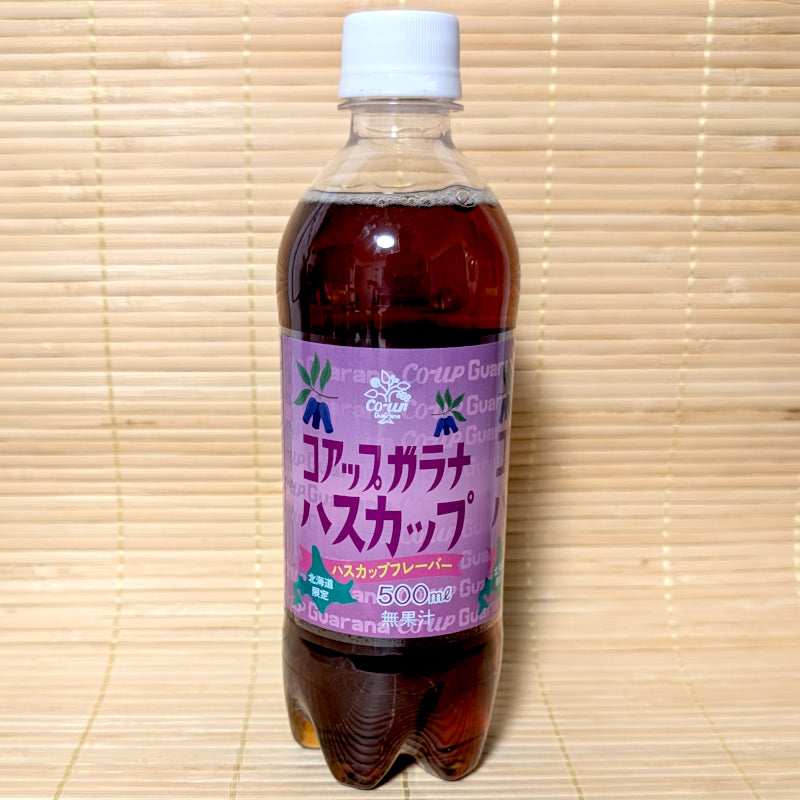 A clear 500ml PET bottle of Co-up Haskap Guarana soda with a purple label on a bamboo mat background.