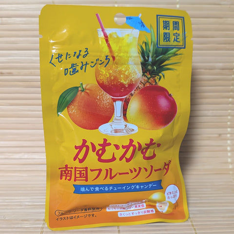 A bright yellow and orange 30g pouch of Kamu Kamu Tropical Fruit Soda candy showing a cocktail glass with an umbrella, fresh mango, pineapple, and citrus fruits on a bamboo mat background.