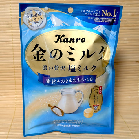 A sky blue and gold pouch of Kanro Kin no Milk Salty Milk candy showing a glass pitcher of fresh milk and a wooden scoop of rock salt on a tatami background.