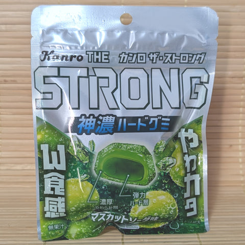 A metallic silver 70g bag of Kanro THE Strong Muscat Soda gummies showing green muscat grapes, cross-section diagrams of the dual-layer gummy, and bold Japanese text on a bamboo mat background.