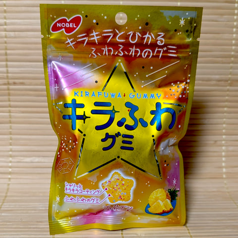 A metallic gold pouch of Nobel Kirafuwa Pineapple gummy on a tatami background featuring a large star and a fluffy star shaped gummy with colorful bits.