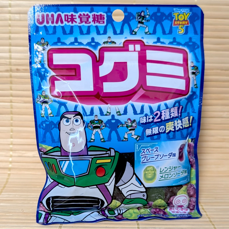 A bright blue 50g resealable pouch of UHA Mikakuto Kogumi gummies showing an animated space ranger character and mini green and purple gummies on a bamboo mat background.