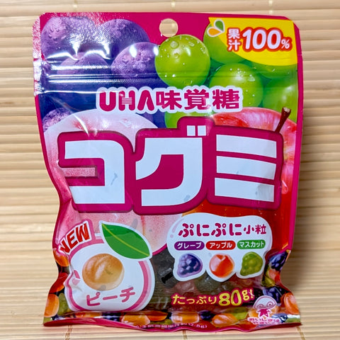 A bright pink and purple 80g bag of UHA Kogumi Gummy Fruit Mix showing real grapes, apples, and peaches against a textured tatami background.
