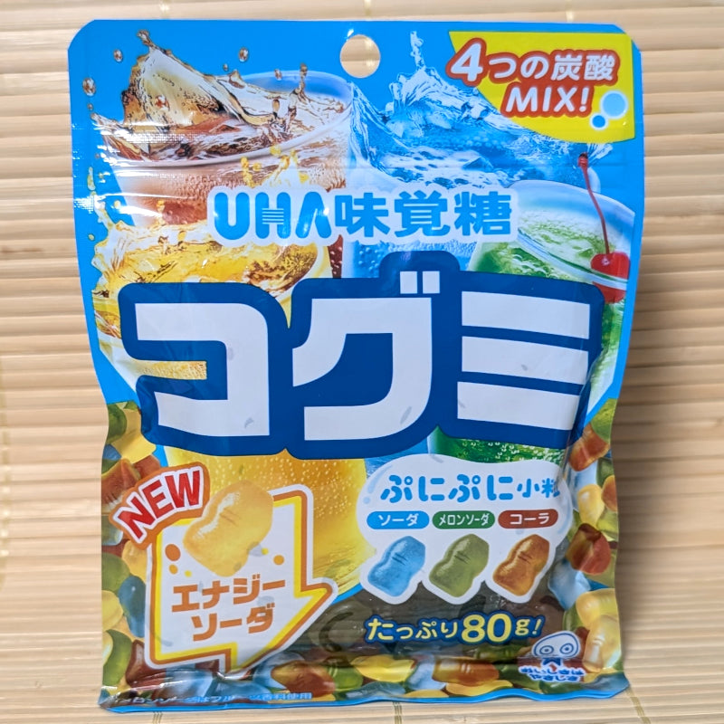 A bright blue 80g bag of UHA Kogumi Gummy Soda Mix showing splashing glasses of cola, ramune, and energy soda against a textured tatami background.