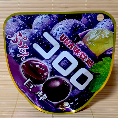 A deep purple and gold 48g resealable pouch of UHA Kororo Kyoho grape gummies displaying fresh Kyoho grapes artwork on a bamboo mat background.