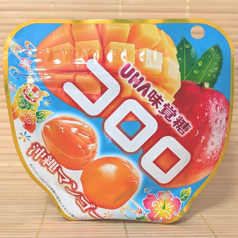 A vibrant blue and yellow 40g pouch of Kororo Okinawa Mango gummies showing sliced mangos, Okinawan Shisa lion art, hibiscus flowers, and round orange gummies on a bamboo mat background.