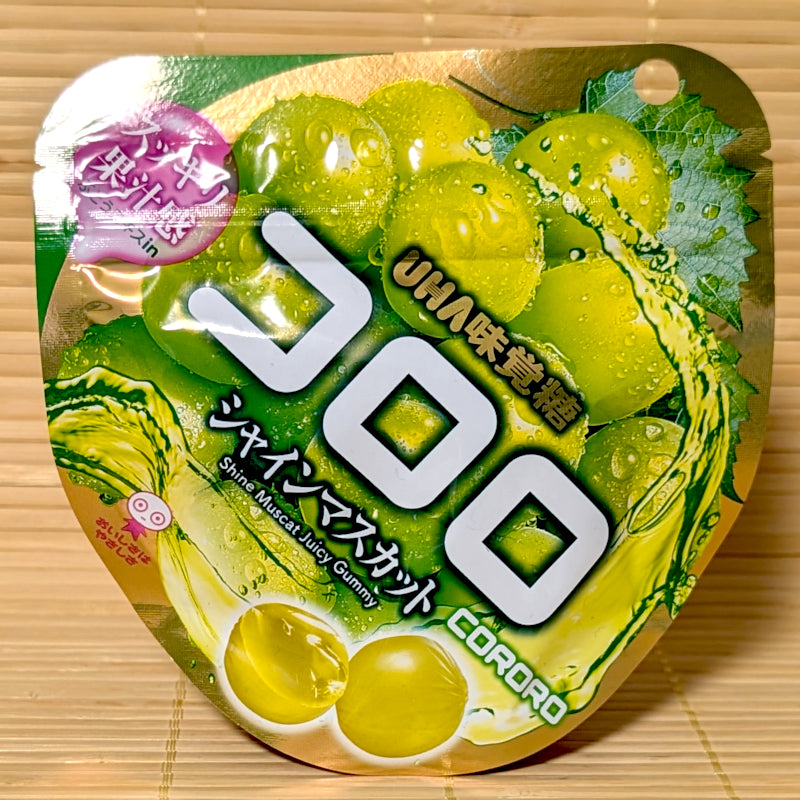 A metallic green and gold pouch of UHA Kororo Shine Muscat Grape Juicy Gummy featuring fresh green grape graphics on a bamboo background.
