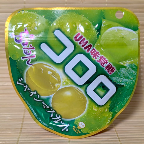 A metallic green and gold 48g resealable pouch of UHA Kororo Shine Muscat grape gummies displaying glistening green grape artwork on a bamboo mat background.