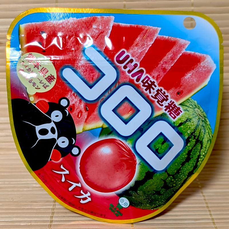 A rounded yellow and blue pouch of UHA Kororo Suika watermelon gummy showing Kumamon, a whole watermelon, and sliced red fruit on a tatami background.