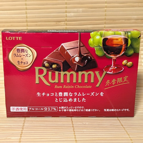 A crimson red box of Lotte Rummy Rum Raisin Chocolate with a glass of rum and grapes on a bamboo background.   