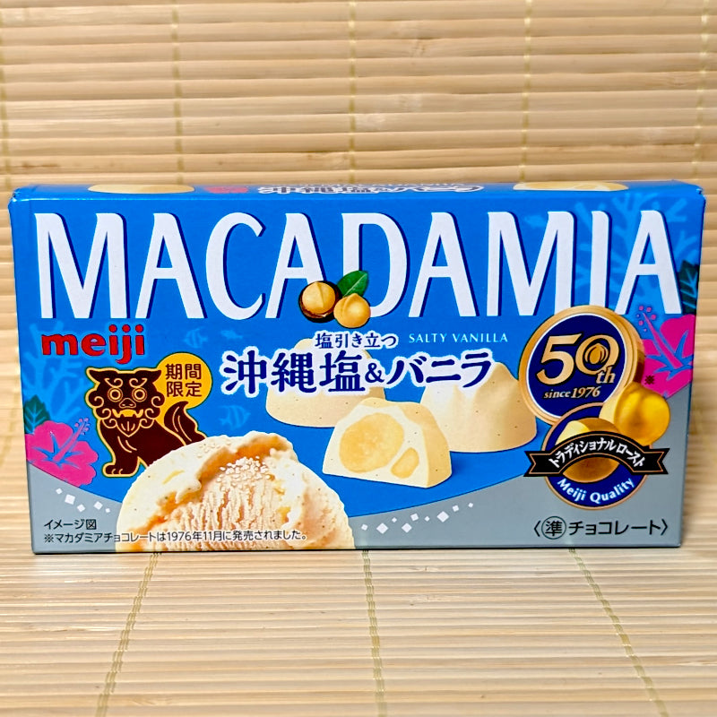 A vibrant blue box of limited edition Meiji Macadamia Okinawa Salty Vanilla chocolate featuring a brown Shisa lion illustration on a tatami background.