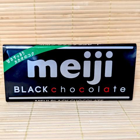 A 50g Meiji Black Chocolate bar wrapped in a sleek black paper label resting on a natural bamboo mat.
