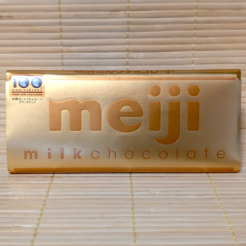 A 50g Meiji Milk Chocolate 100th Anniversary bar wrapped in a commemorative gold paper sleeve with gold lettering, resting on a natural bamboo mat.