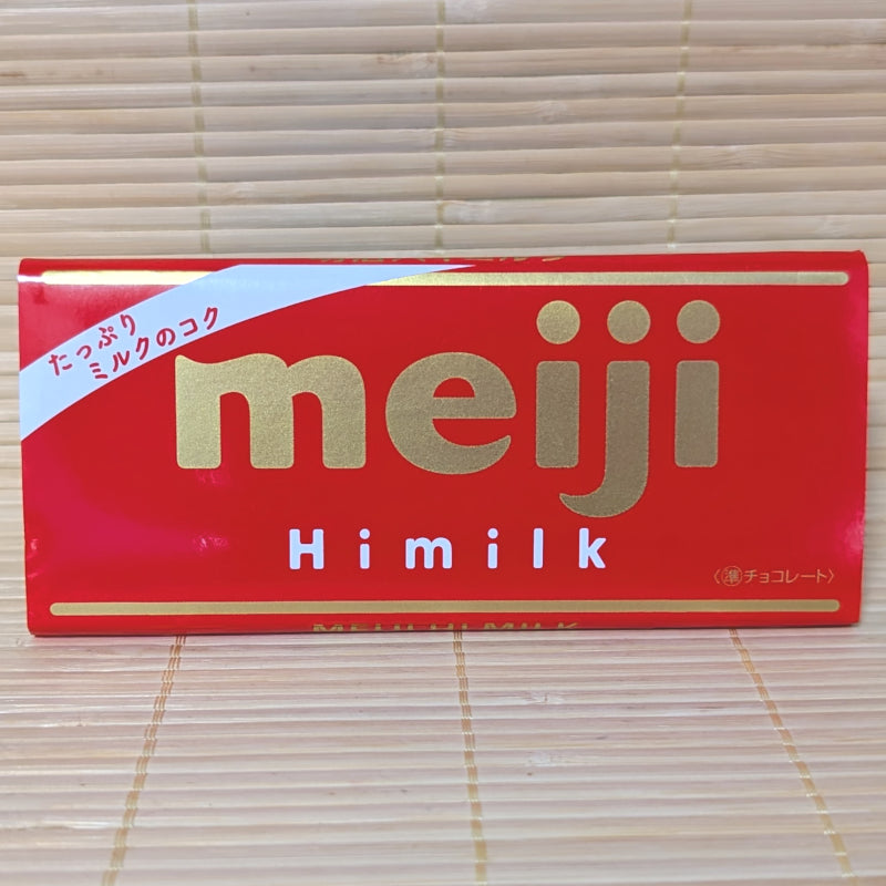 A 50g Meiji Hi-Milk Chocolate bar wrapped in a red paper label with gold lettering, resting on a natural bamboo mat.