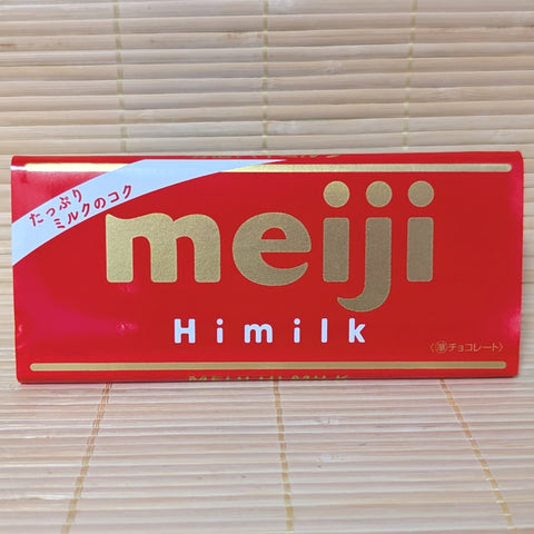 A 50g Meiji Hi-Milk Chocolate bar wrapped in a red paper label with gold lettering, resting on a natural bamboo mat.