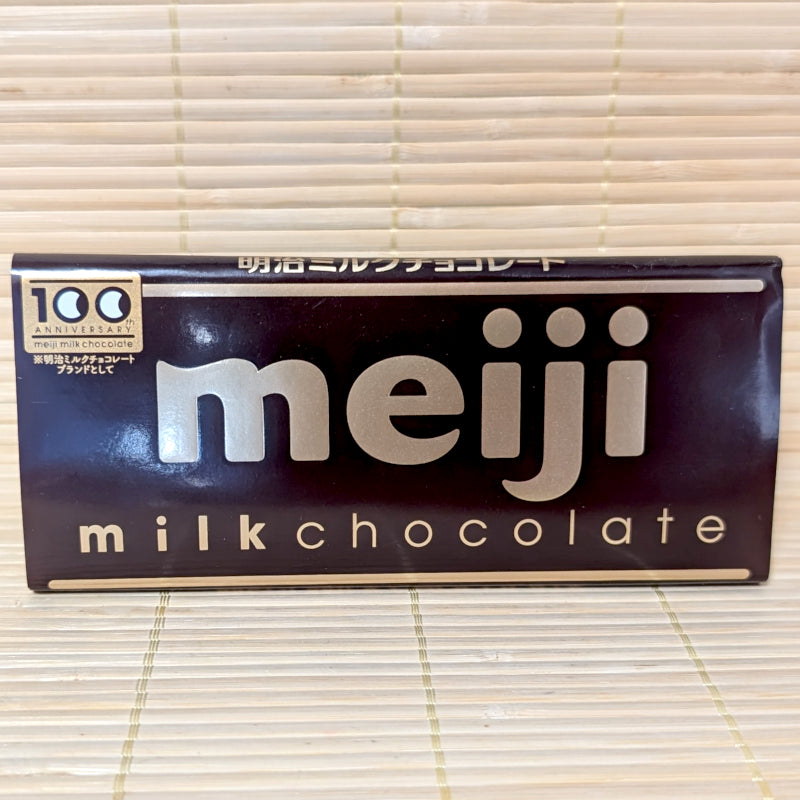A 50g Meiji Milk Chocolate bar wrapped in a classic brown paper label with gold lettering, resting on a natural bamboo mat.