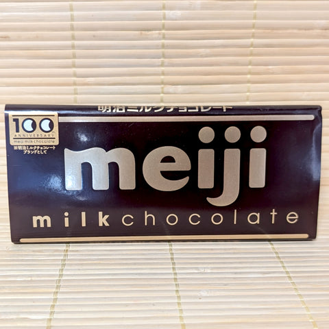 A 50g Meiji Milk Chocolate bar wrapped in a classic brown paper label with gold lettering, resting on a natural bamboo mat.