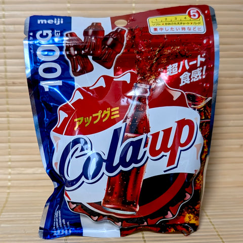 A metallic red and blue 100g resealable bag of Meiji Cola Up gummies showing bottle shapes and an exploding soda graphics on a bamboo mat background.