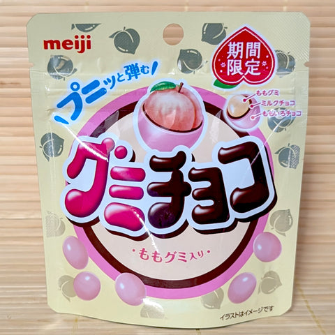 A cream and pink 40g pouch of Meiji Gummy Choco Peach showing a fresh white peach, cross-section diagram of the triple-layer chocolate gummy ball, and pink candy pieces on a bamboo mat background.