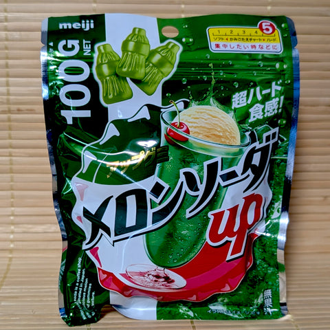 A metallic deep green 100g resealable bag of Meiji Melon Soda Up gummies showing a melon soda float with vanilla ice cream and bottle shapes on a bamboo mat background.