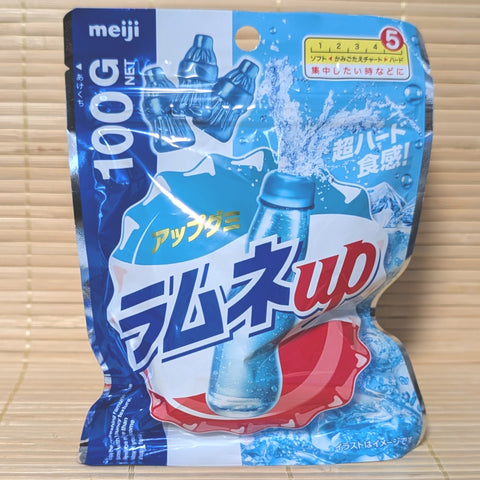 A metallic blue 100g resealable bag of Meiji Ramune Up gummies showing soda bottle shapes and splashing blue liquid on a bamboo mat background.