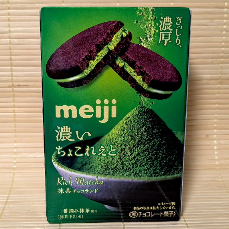 A deep green box of Meiji Rich Matcha Chocolate Sandwich Cookies showing dark cocoa cookies with vivid green matcha filling and a bowl of powdered matcha on a bamboo mat background.