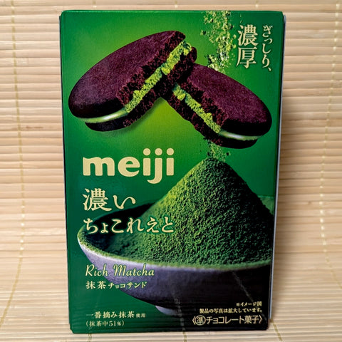 A deep green box of Meiji Rich Matcha Chocolate Sandwich Cookies showing dark cocoa cookies with vivid green matcha filling and a bowl of powdered matcha on a bamboo mat background.