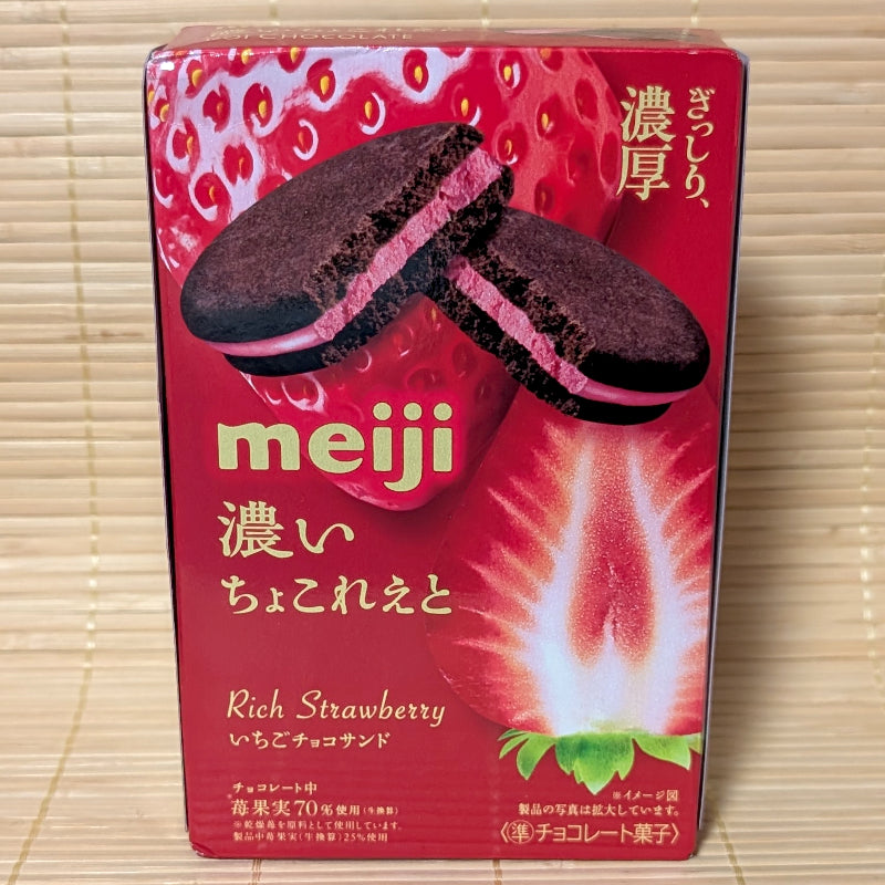 A vibrant red box of Meiji Rich Strawberry Chocolate Sandwich Cookies showing dark cocoa cookies with pink strawberry filling and fresh strawberries on a bamboo mat background.