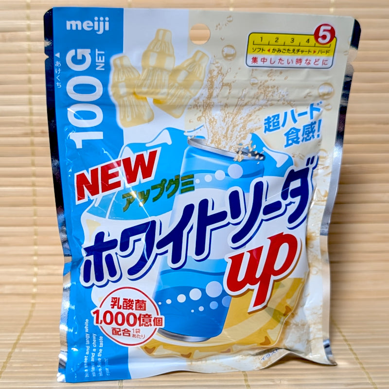 A metallic blue and white 100g resealable bag of Meiji White Soda Up gummies showing a bubbly soda can, a red lactic acid circle logo, and white bottle shapes on a bamboo mat background.