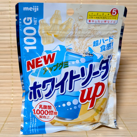 A metallic blue and white 100g resealable bag of Meiji White Soda Up gummies showing a bubbly soda can, a red lactic acid circle logo, and white bottle shapes on a bamboo mat background.