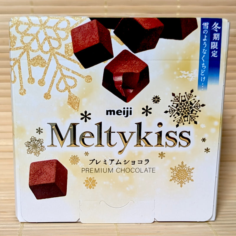 A white and gold 52g box of Meiji Meltykiss Premium Chocolate showing snowflake designs and cocoa-dusted chocolate cubes on a bamboo background.   
