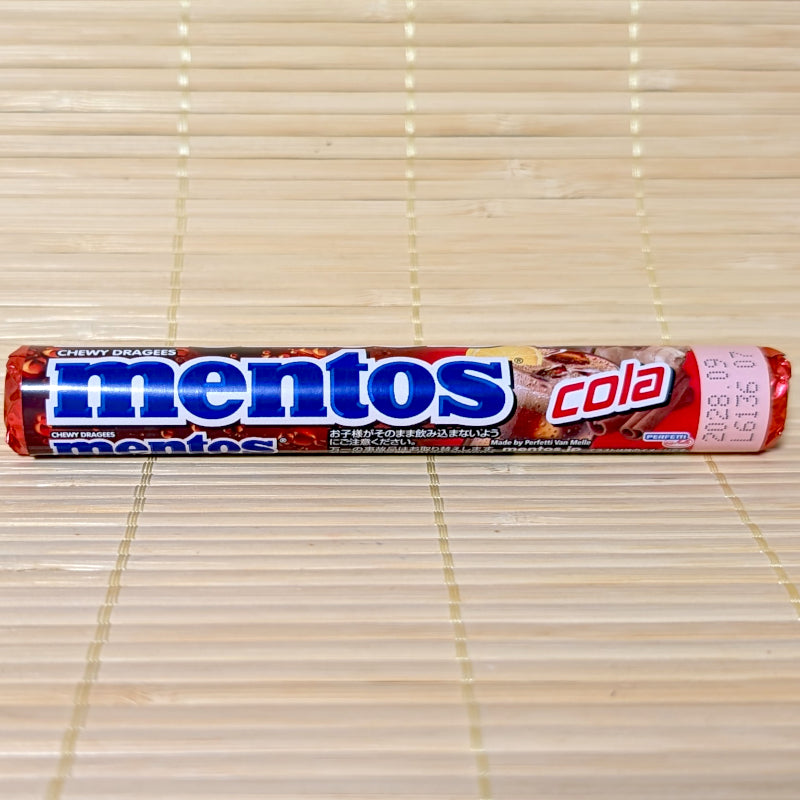 A single roll of Mentos Cola chewy candy featuring a deep red and brown wrapper with the classic blue Mentos logo, displayed horizontally on a tatami mat background.