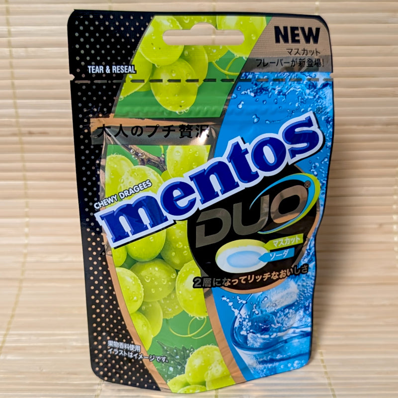 A bright green and blue resealable Mentos pouch showing a cross-section of a green grape and soda-flavored candy. Displayed on a tatami surface.