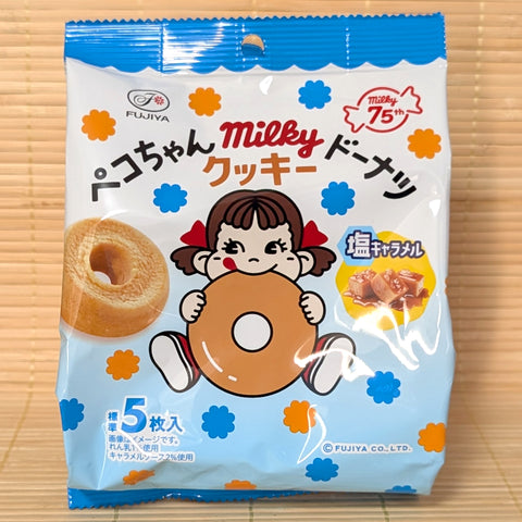A light blue and white 60g pouch of Fujiya Peko-chan Milky Cookie Donuts Salted Caramel showing Peko-chan holding a donut, caramel cubes, and a soft mini cake ring on a bamboo mat background.