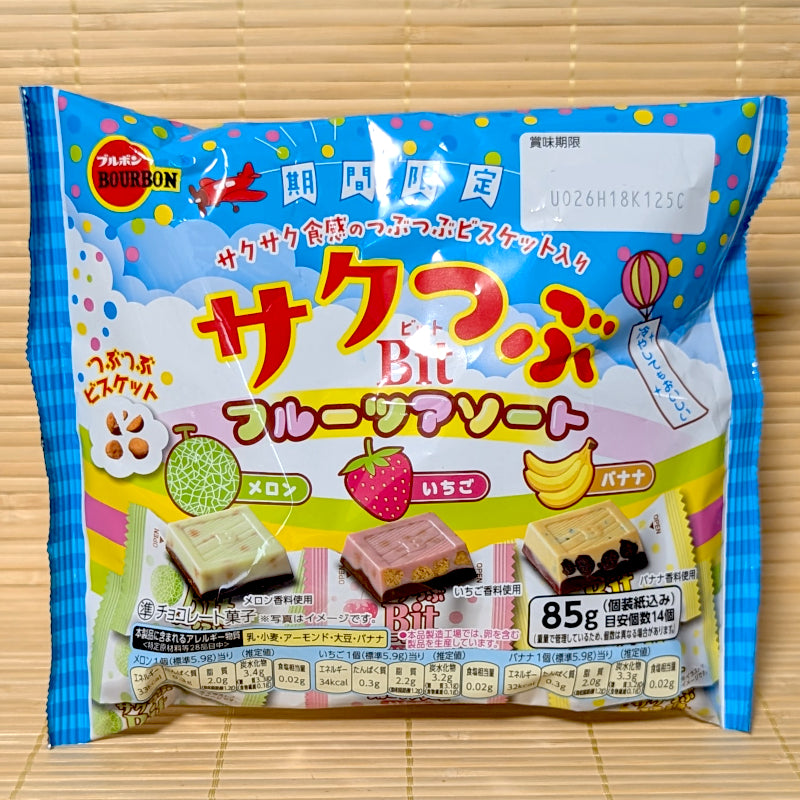 A bright blue and yellow 85g bag of Bourbon Mini Bit fruit chocolate assortment showing cartoon graphics of a plane, a hot air balloon, fresh melon, strawberry, and banana next to cross-section diagrams of the crispy cookie chocolates on a bamboo mat background.