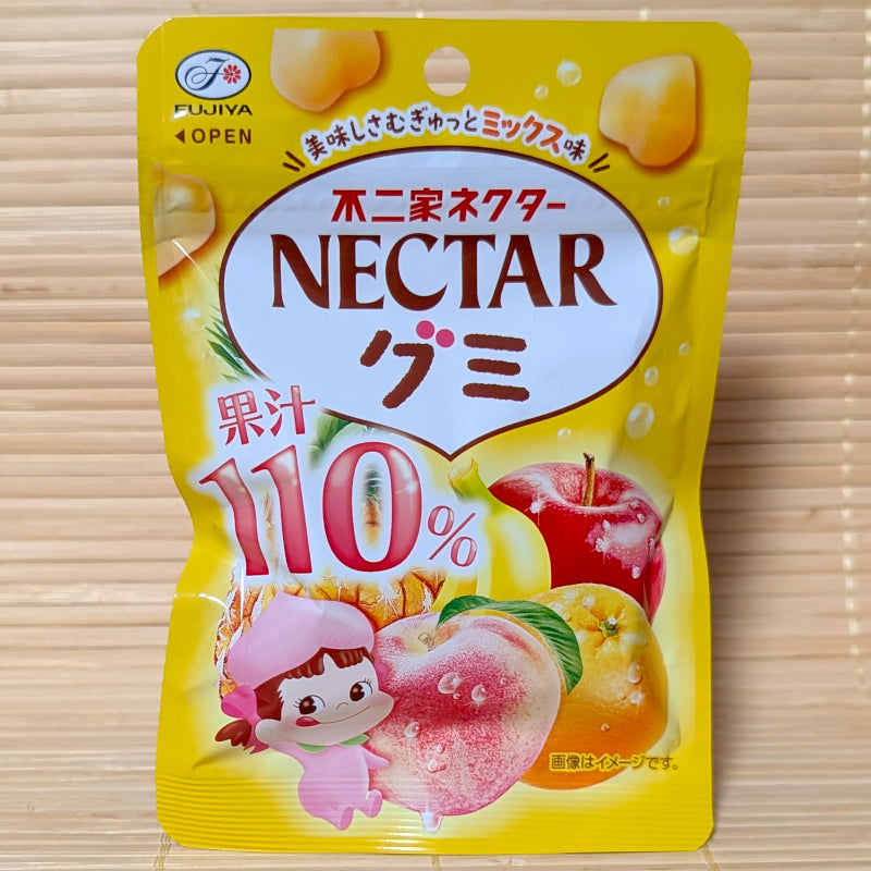 A bright yellow 54g resealable pouch of Fujiya Nectar 110% Mixed Fruit gummies with peach, pineapple, apple, banana, and orange graphics on a bamboo mat background.